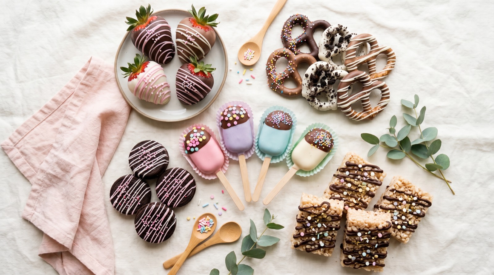 An assortment of chocolate-covered strawberries, pretzels, cake pops, drizzled Oreos and rice krispie treats arranged on a soft cream cloth
